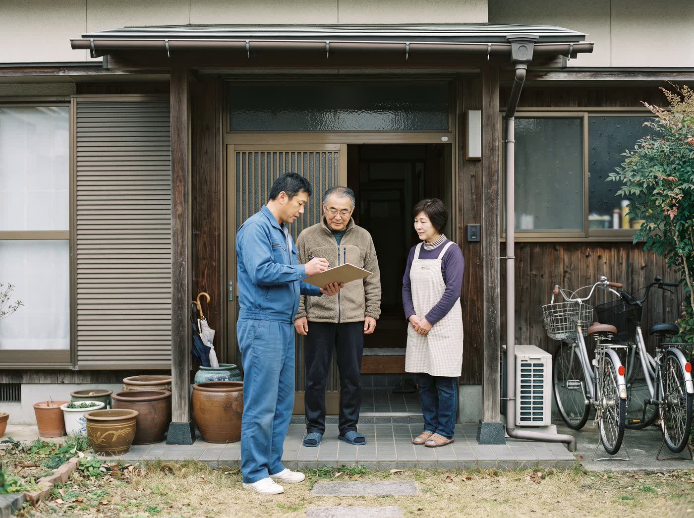 住宅の玄関先で、作業着姿のスタッフが住人の夫婦に点検結果を説明している様子